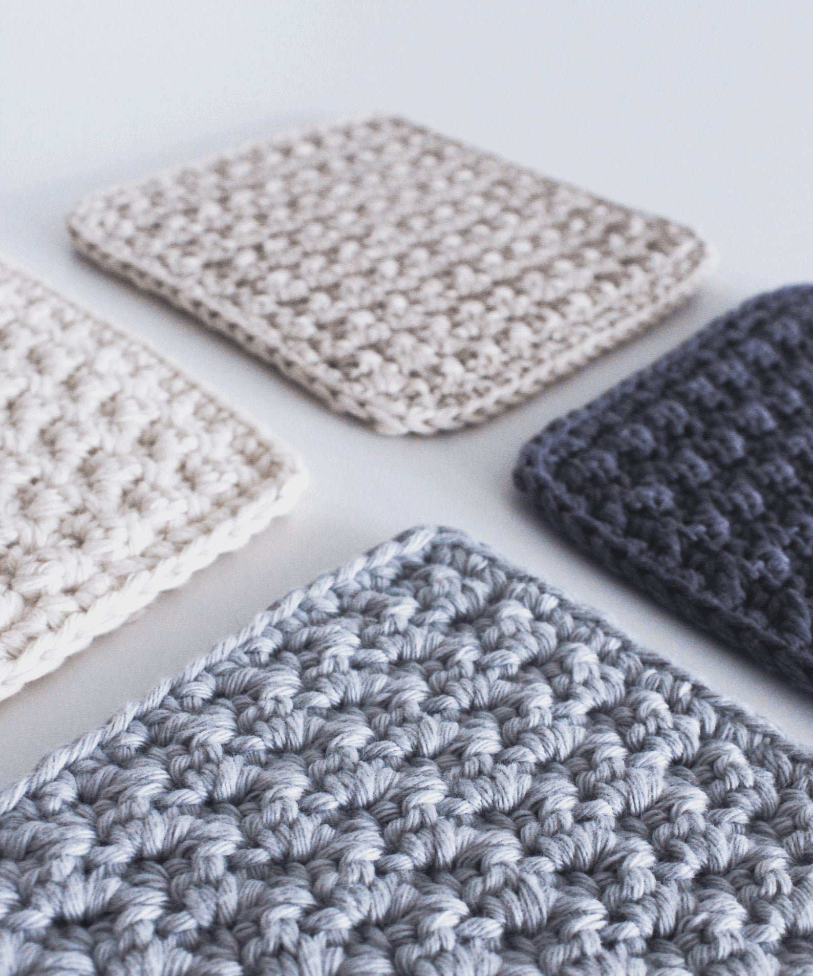 Four textured square crochet coasters in complementary colours; pale blue, charcoal, cream and beige.