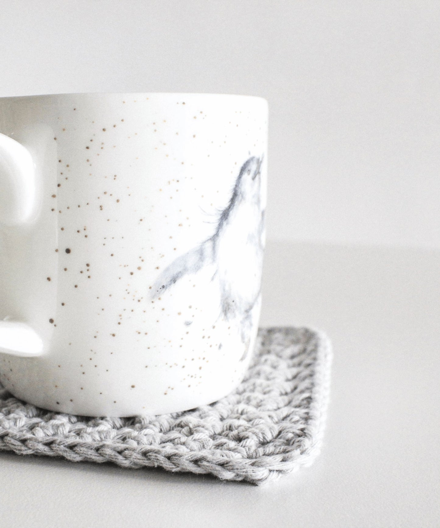 A grey crochet coaster sits on a white table with a white mug on it. The mug has a hand-drawn baby penguin illustration.