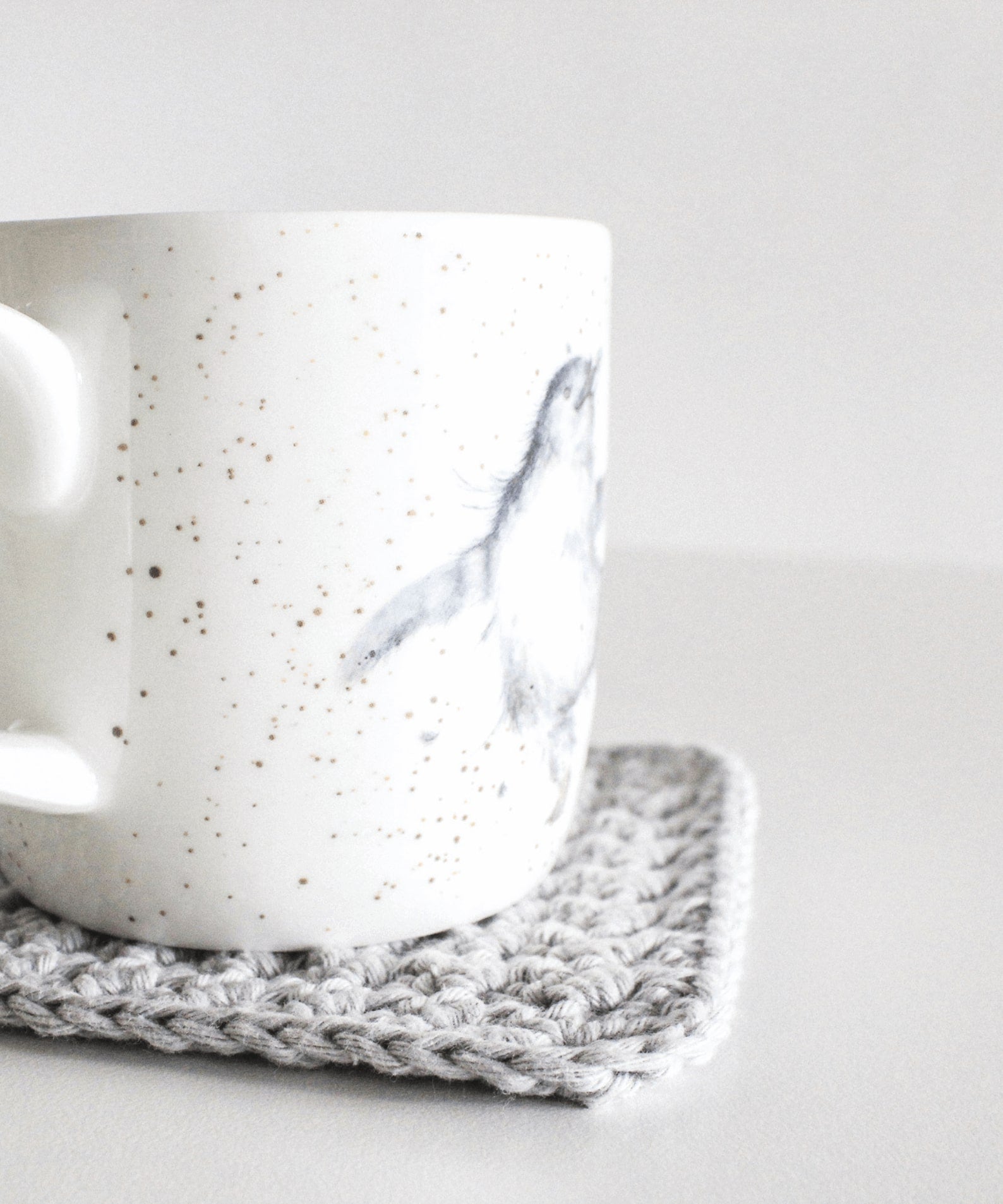 A grey crochet coaster sits on a white table with a white mug on it. The mug has a hand-drawn baby penguin illustration.