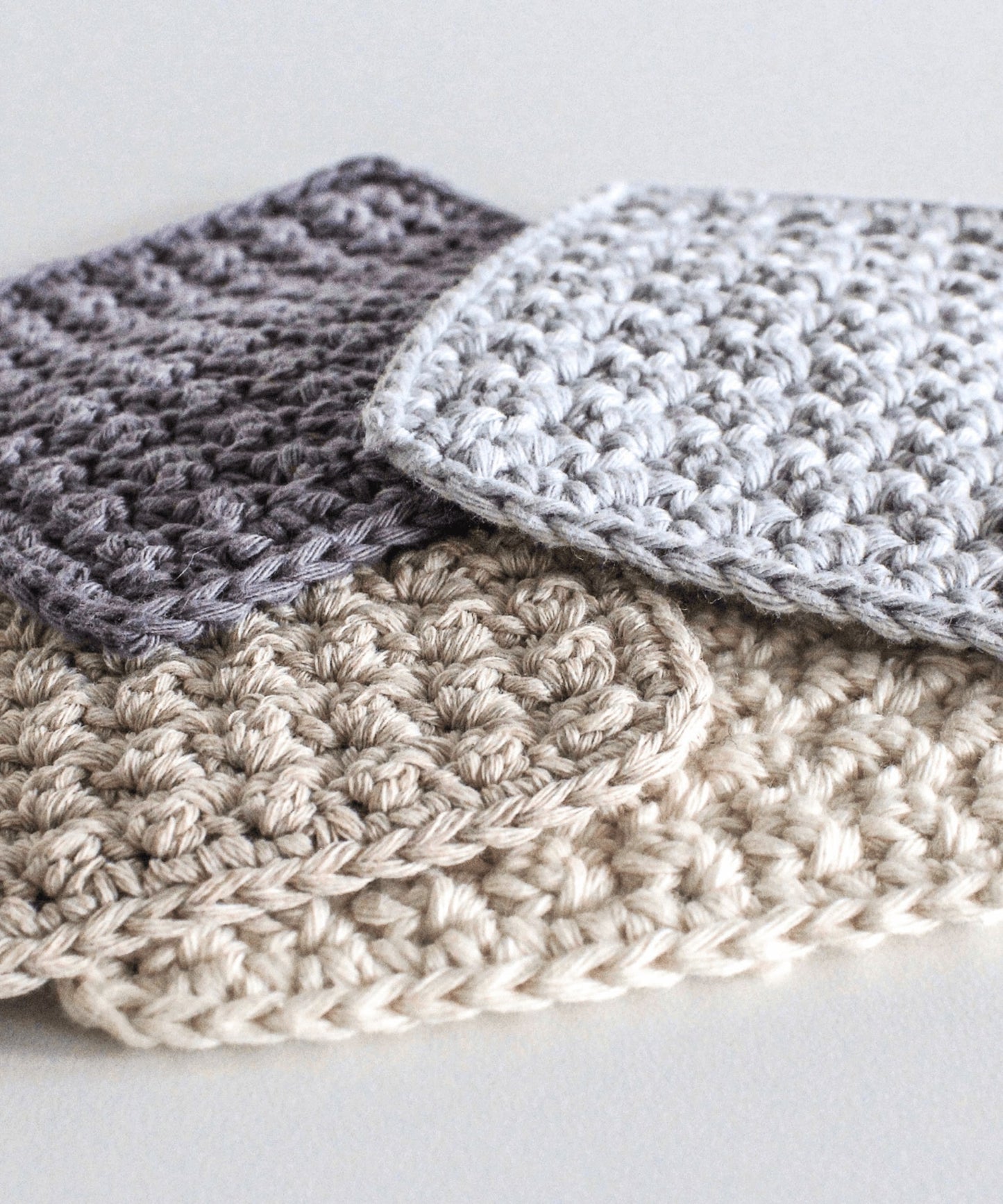 Four square crochet coaster are stacked loosely in a pile. They are in complementary colours; charcoal, pale blue, cream and beige.