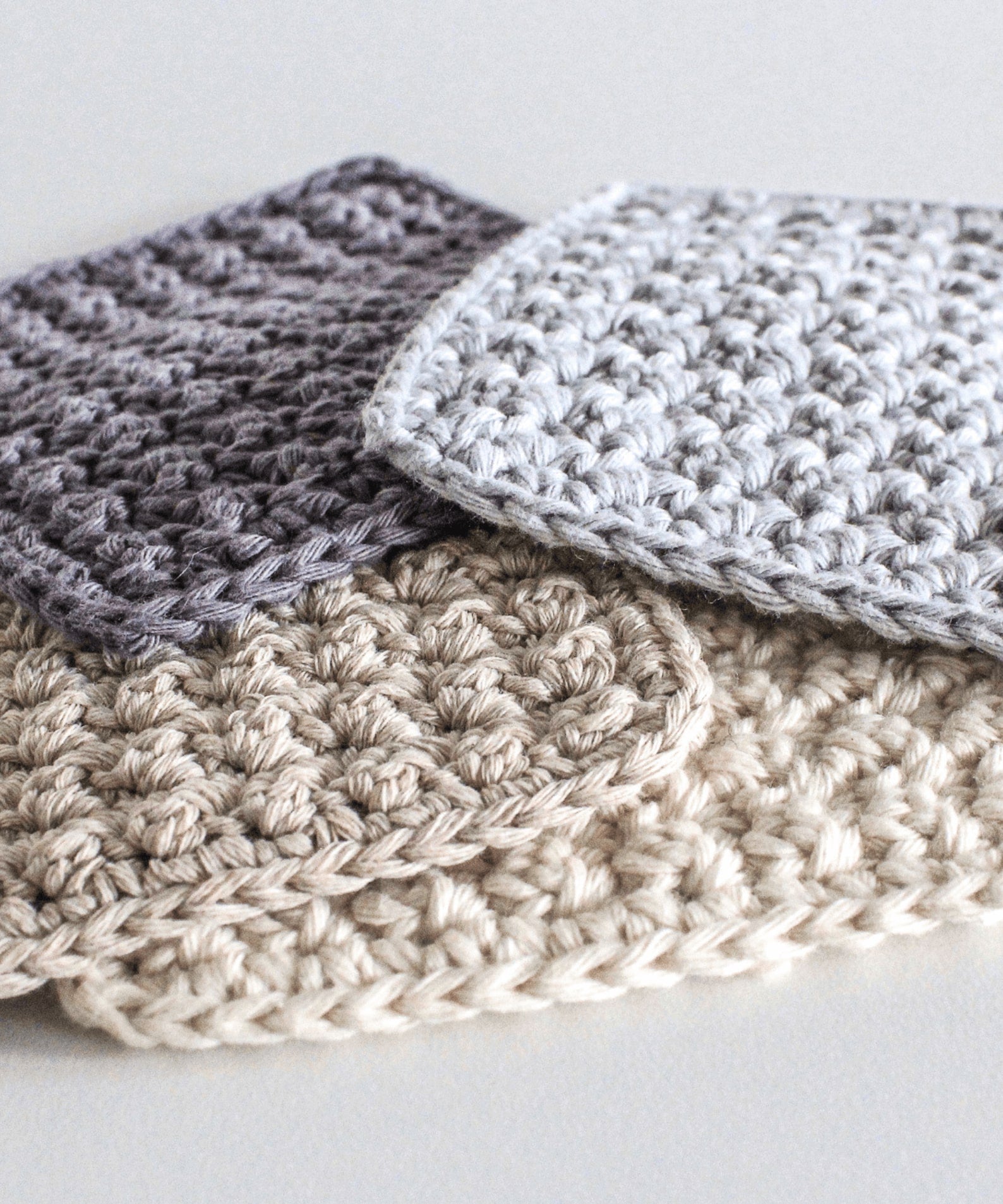 Four square crochet coaster are stacked loosely in a pile. They are in complementary colours; charcoal, pale blue, cream and beige.