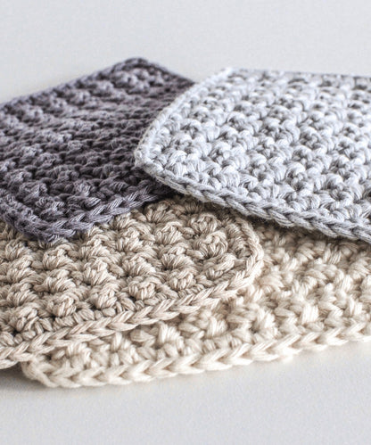 Four square crochet coaster are stacked loosely in a pile. They are in complementary colours; charcoal, pale blue, cream and beige.